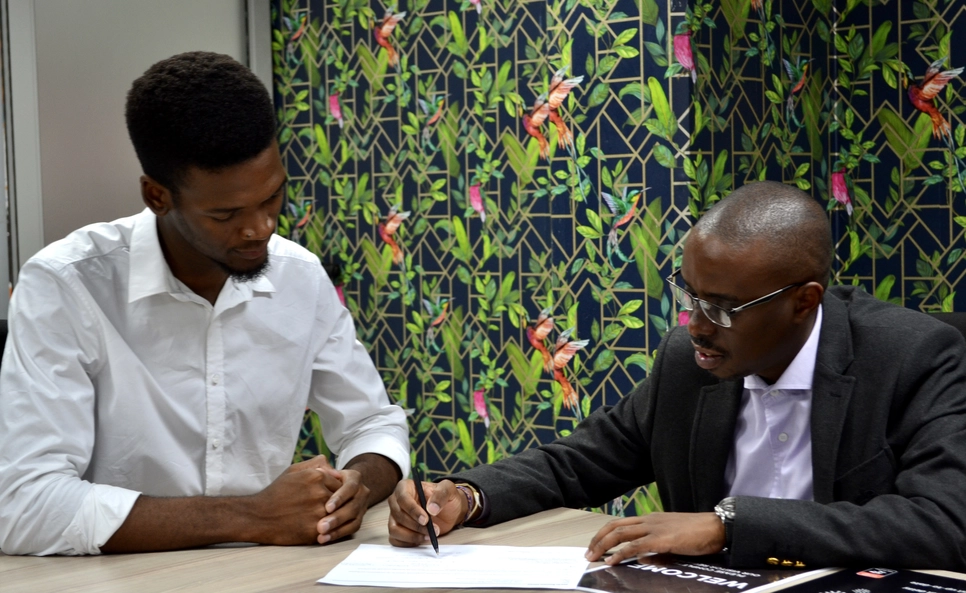 Architect Siyanda Mthembu from Durban in a focused meeting with a client, signing documents on the table. They are in an office with an eye-catching tropical bird and foliage-patterned backdrop.