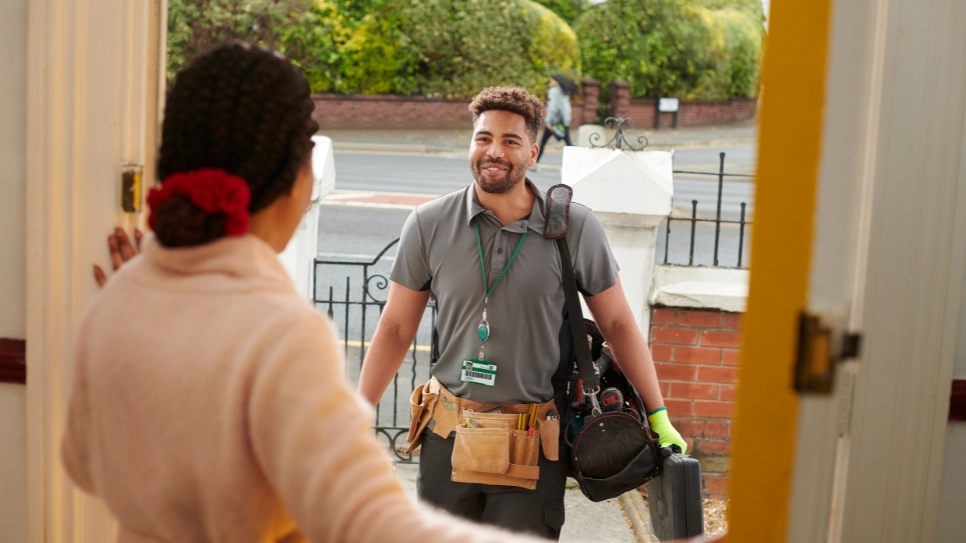 A smiling tradesman with a badge and a tool belt arrives at a customer's open front door, greeted by a woman with a welcoming gesture. The scene is set in a residential area during daytime.