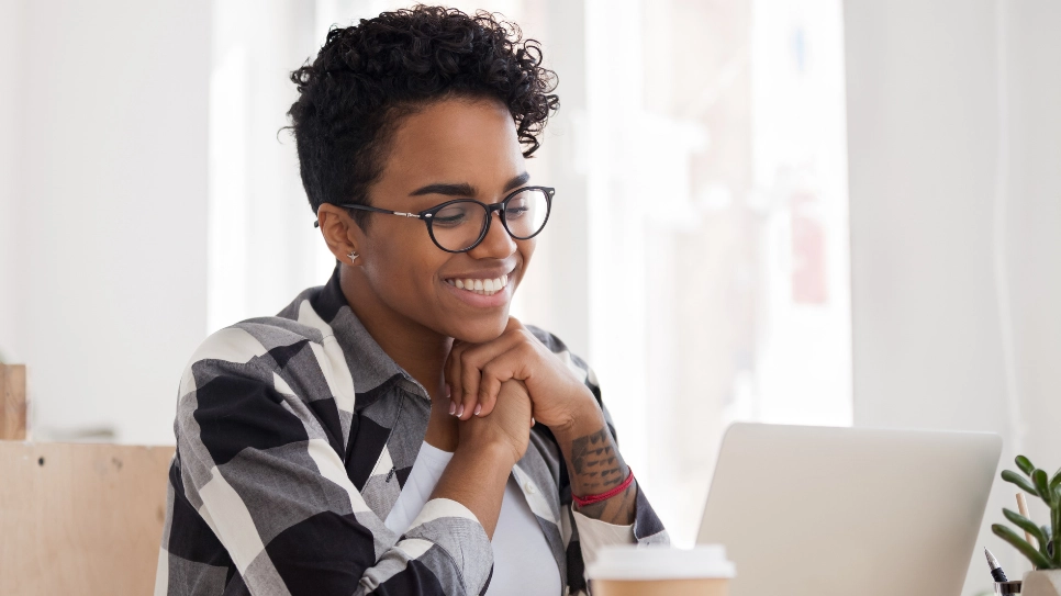 A cheerful young woman with short curly hair and glasses smiling while looking at her new businbess website at her laptop, seated in a bright office space. She's wearing a black and white checked shirt and has a red wristband, suggesting a casual yet professional setting.