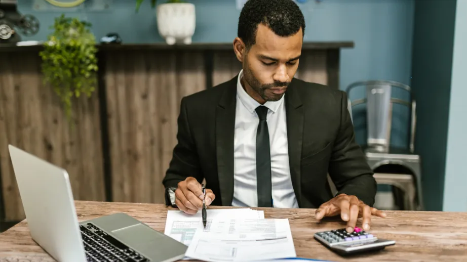 Professional in a suit reviewing tax documents at a desk, using a calculator beside a laptop in an office.