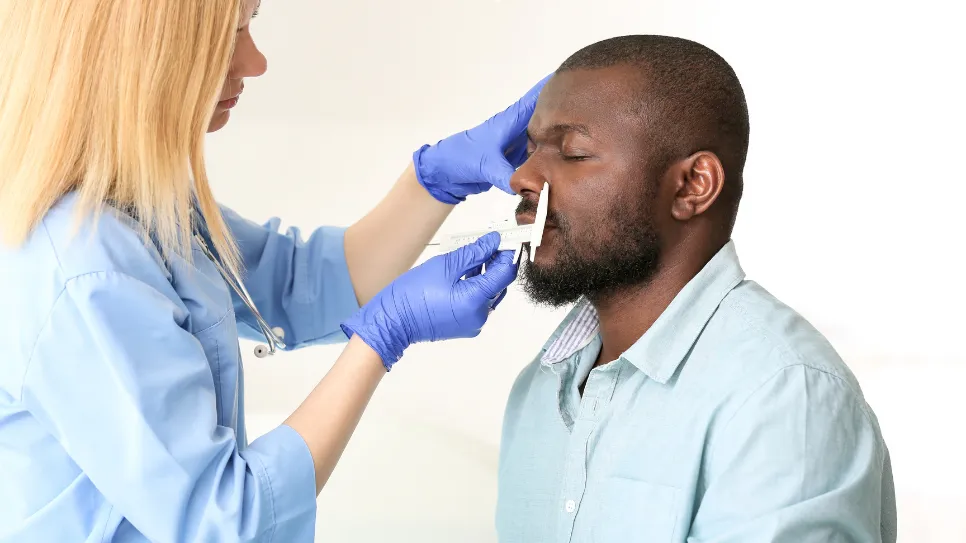 Medical professional measuring a patient's nose during a pre-operative consultation, assessing facial dimensions for surgery planning.