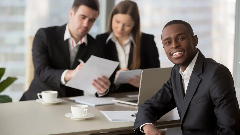 A confident professional sits in the foreground while two HR team members review documents in a modern office setting.