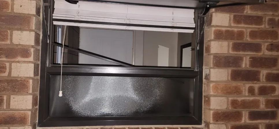 A window with a broken hinge mechanism, causing it to remain partially open, set in a brick wall.