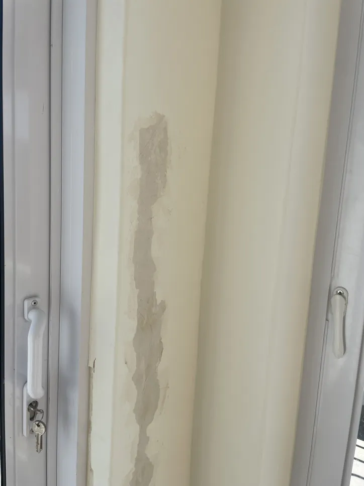 Peeling paint on a wall near a white window frame, indicating possible moisture issues.
