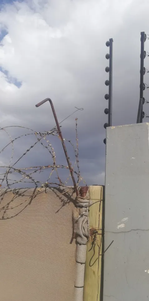 Barbed wire and electric fence atop a concrete wall under cloudy skies, enhancing security measures.