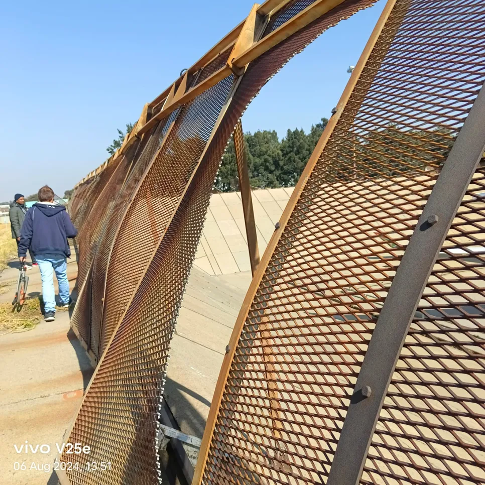 Damaged metal mesh fence (23176) Bent metal mesh fence along a pathway with two people walking nearby, indicating potential repair needs.