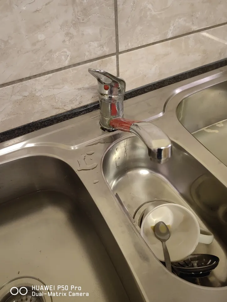 Leaking kitchen faucet with visible water droplets on stainless steel sink and unwashed dishes.