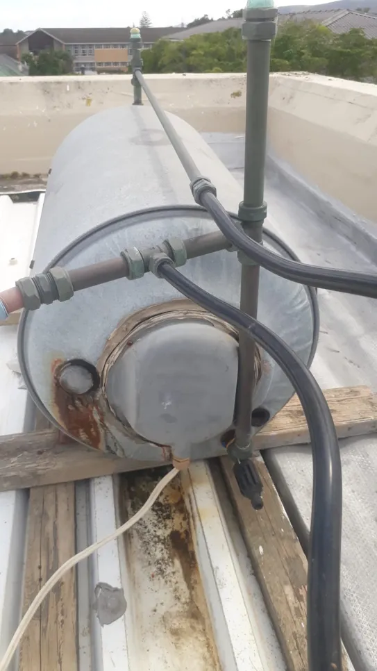 Rooftop water heater tank showing signs of rust and corrosion, with connected pipes and fittings.