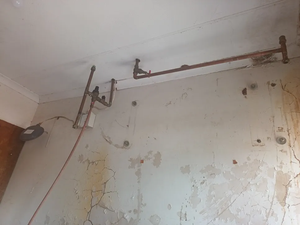 Exposed plumbing pipes on a wall with peeling paint and visible water damage signs.