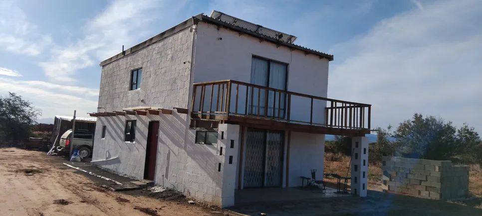 Unfinished two-story concrete block house with a wooden balcony, located in a rural area under a clear sky.