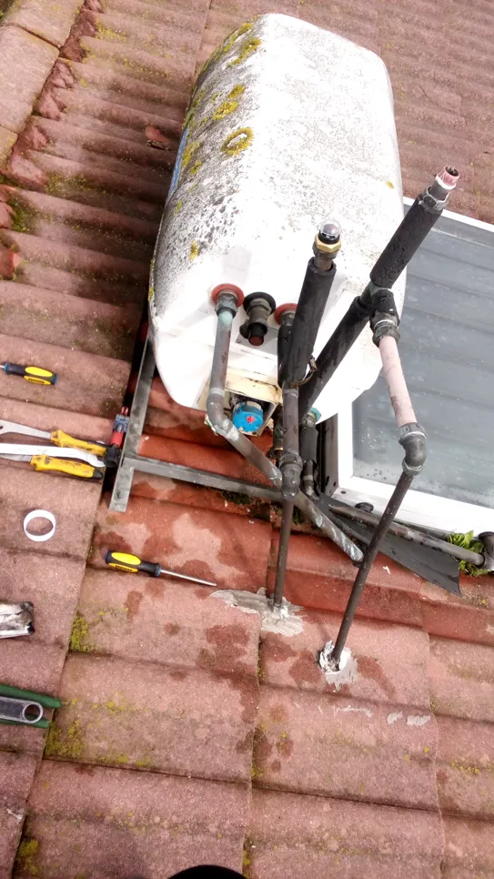 Rooftop solar water heater installation (19254) Old solar water heater on a tiled roof with visible pipes and tools scattered around.