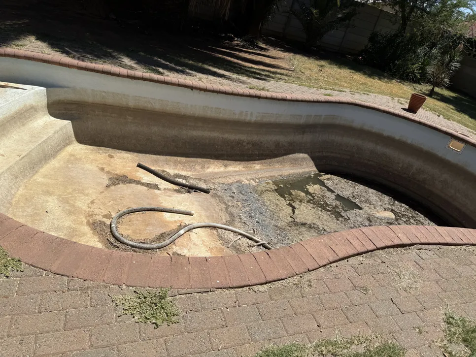 An empty swimming pool with cracked surfaces and debris, surrounded by a brick patio in a backyard setting.