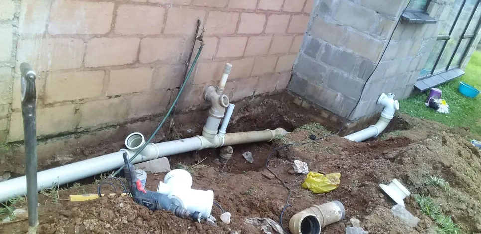 Outdoor plumbing pipes installed along a house foundation with tools and soil around.