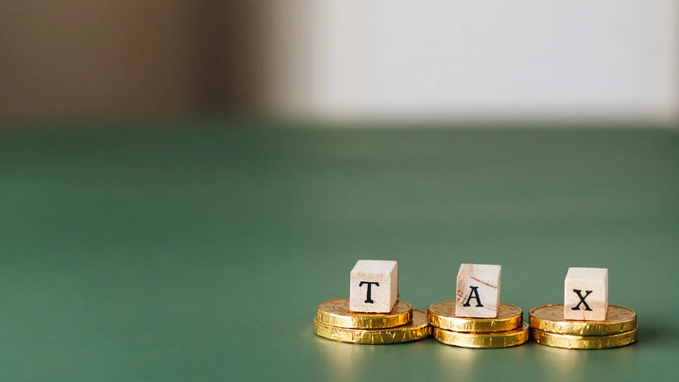 Wooden blocks spelling the word "TAX" resting on three separate stacks of gold coins. The arrangement sits on a solid green surface with a neutral background.