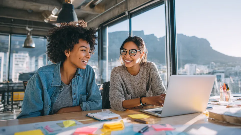 Two smiling female colleagues work together on a laptop at a desk in a modern office with large windows overlooking Table Mountain and the city of Cape Town.