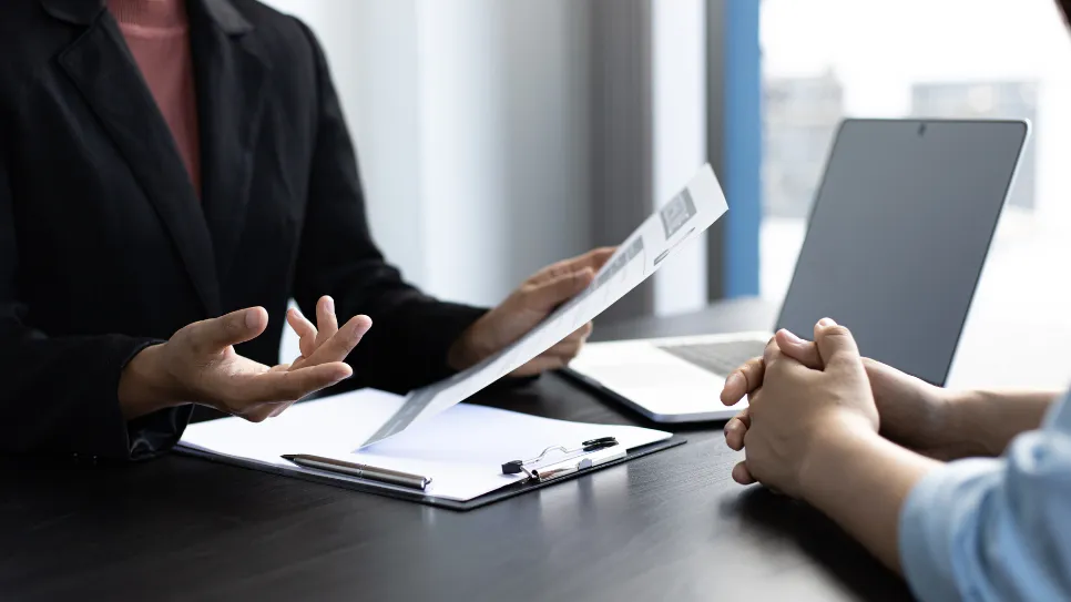 During a job interview, a human resources manager in a black blazer gestures while discussing the candidate's resume, with the candidate's clasped hands visible across the desk.