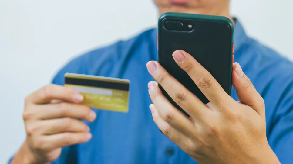 A person in a blue shirt holds a smartphone in one hand and a credit card in the other, appearing to make an online purchase or payment.