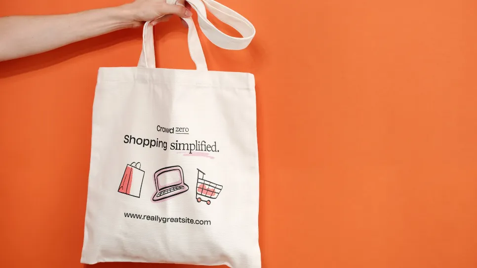 A hand holds up a white canvas tote bag against a bright orange wall; the bag is printed with the words "crowd zero, shopping simplified" and illustrations of a shopping bag, a laptop, and a shopping cart.