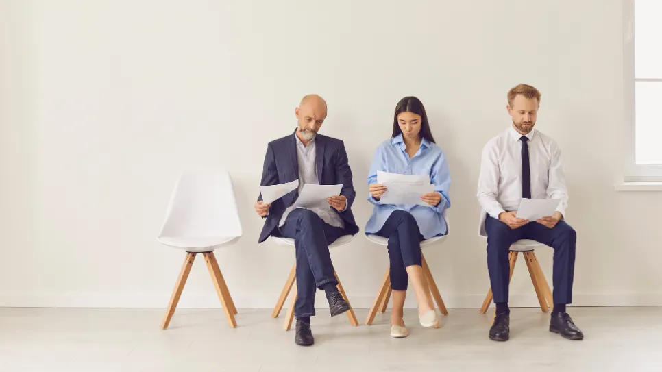 Three  job candidates, an older man in a blazer, a young woman, and a younger man in a tie, sit in a line reviewing documents while waiting for their interviews in a minimalist waiting area.