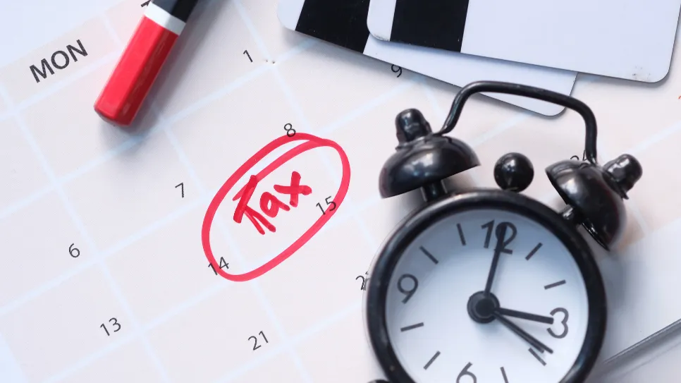 A close-up of a calendar with the word "Tax" written in red ink and circled on the 14th of the month. A black alarm clock, a red marker, and some cards are also on the calendar, signifying a tax deadline.