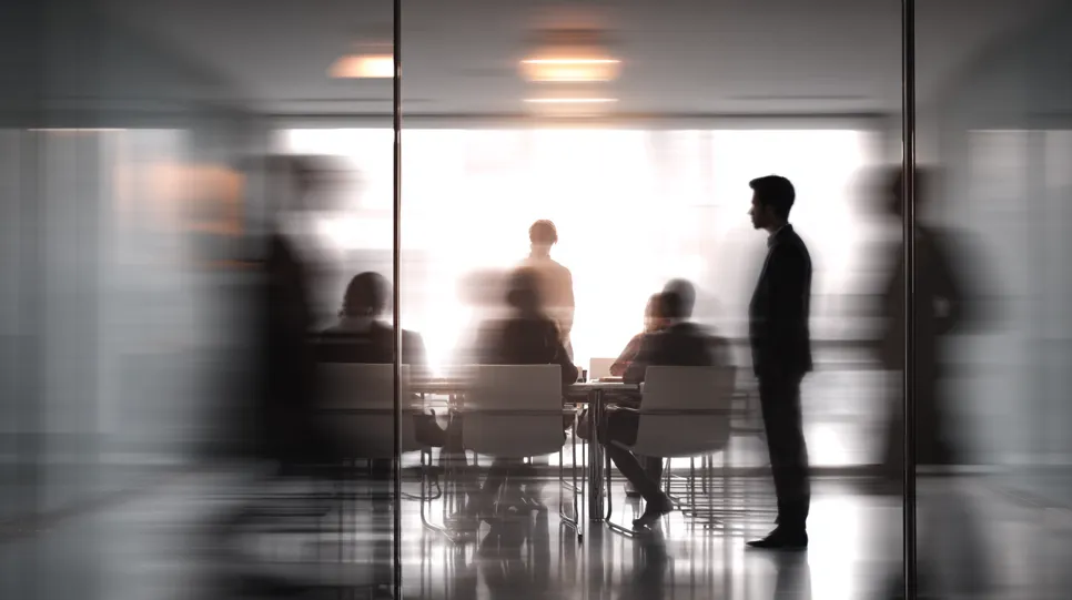 An abstract, motion-blurred view through a glass wall into a conference room where a disciplinary hearing is in progress. The silhouetted figures and blurred motion suggest the confidential and serious nature of the formal meeting.