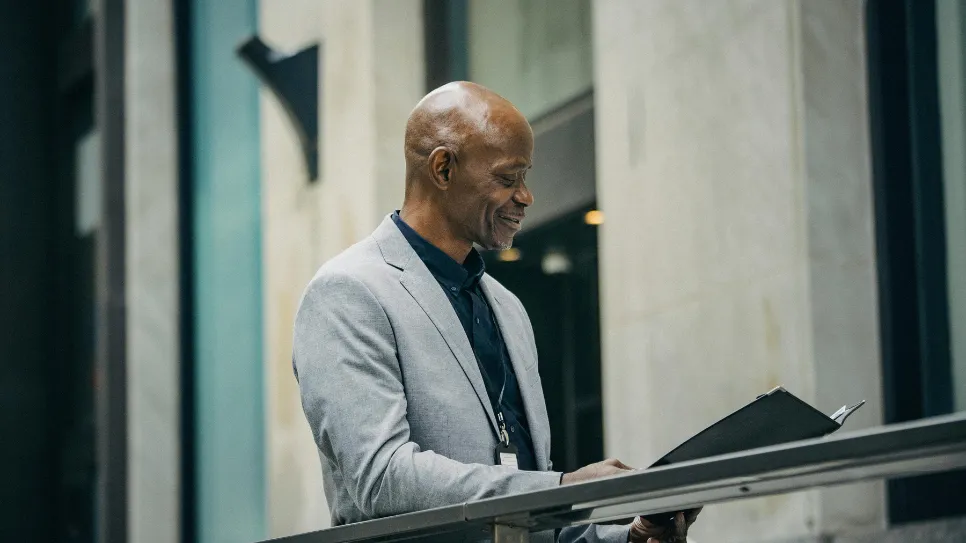 A smiling, bald Black professional in a grey blazer stands in a modern office, looking pleased as he reviews documents in a folder, symbolizing successful human resources management and employee satisfaction.