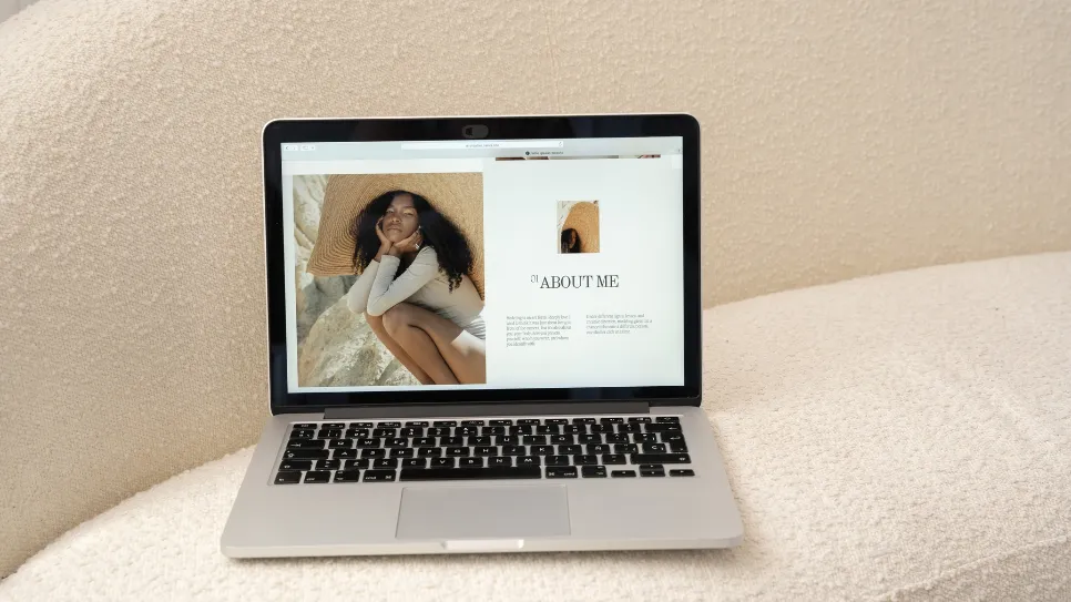 A silver laptop resting on a cream-colored, textured bouclé couch. The laptop screen displays a personal website's 'About Me' page. The page features a large photo of a Black woman with long, dark hair wearing a large straw hat, and the text is in a minimalist font. A black privacy cover is visible over the laptop's webcam.