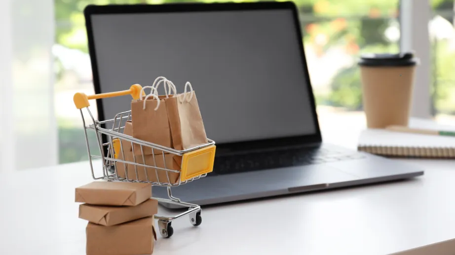 A miniature shopping cart filled with small paper bags placed in front of a laptop on a desk, symbolizing online shopping and digital retail.