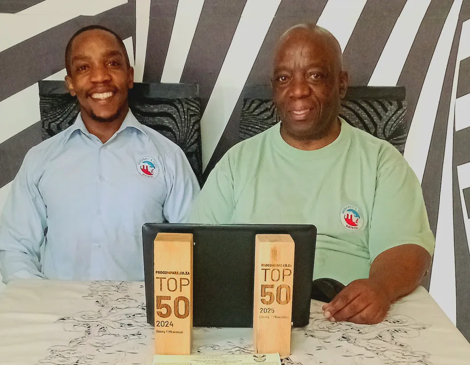 Danny T Mkwanazi and a colleague share a victorious moment behind their pair of Procompare.co.za TOP 50 trophies. The 2024 and 2025 awards sit side-by-side on the table, showcasing a powerful two-year legacy of elite electrical service and community trust.