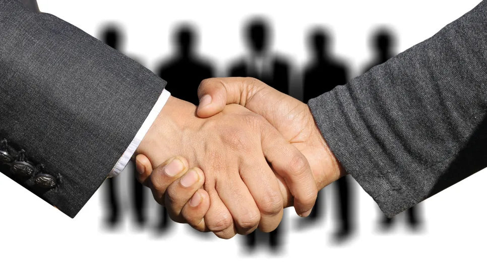 Close-up photograph of two individuals in business attire shaking hands, with blurred silhouettes of a team of people standing in the background.