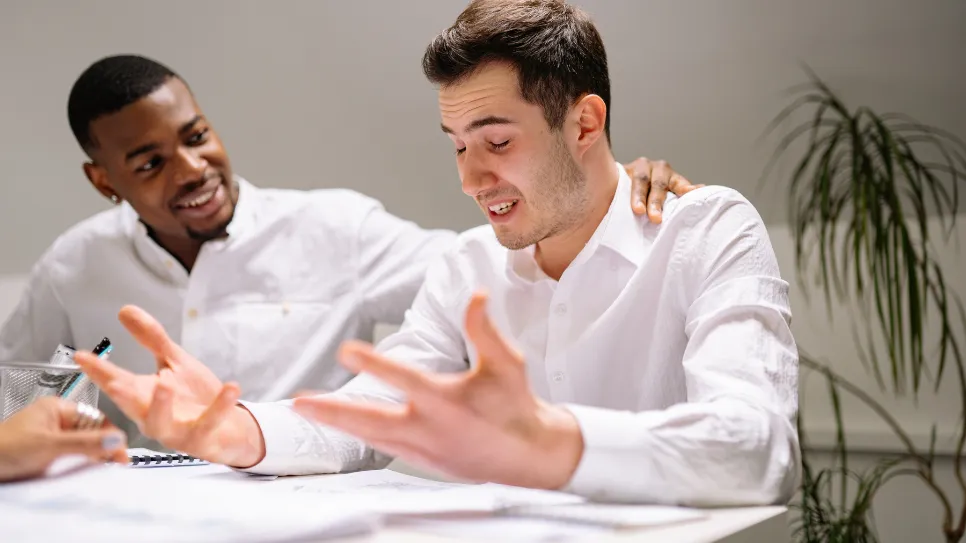 Photograph of a man in a white shirt expressing frustration with raised hands during a meeting while a colleague supportively places a hand on his shoulder and smiles.